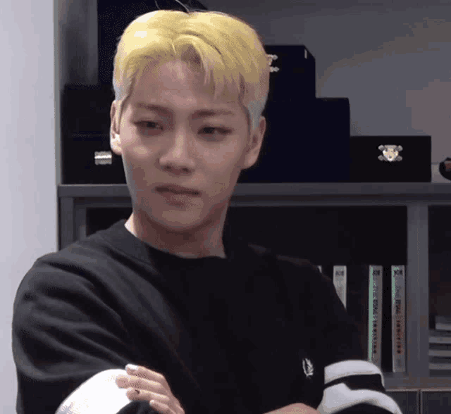 a young man with yellow hair is standing with his arms crossed in front of a bookshelf