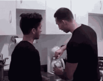 two men are standing next to each other in a kitchen . one of the men is pouring milk into the other 's glass .