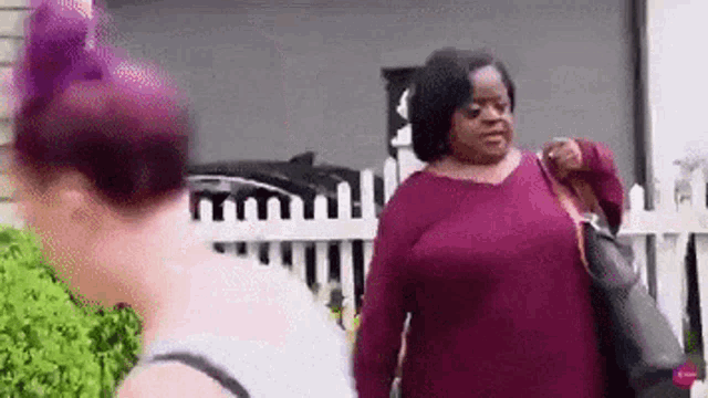 two women are standing next to each other in front of a white picket fence and talking to each other .