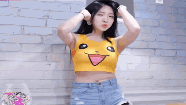 a woman wearing a yellow crop top and blue shorts is dancing in front of a brick wall .
