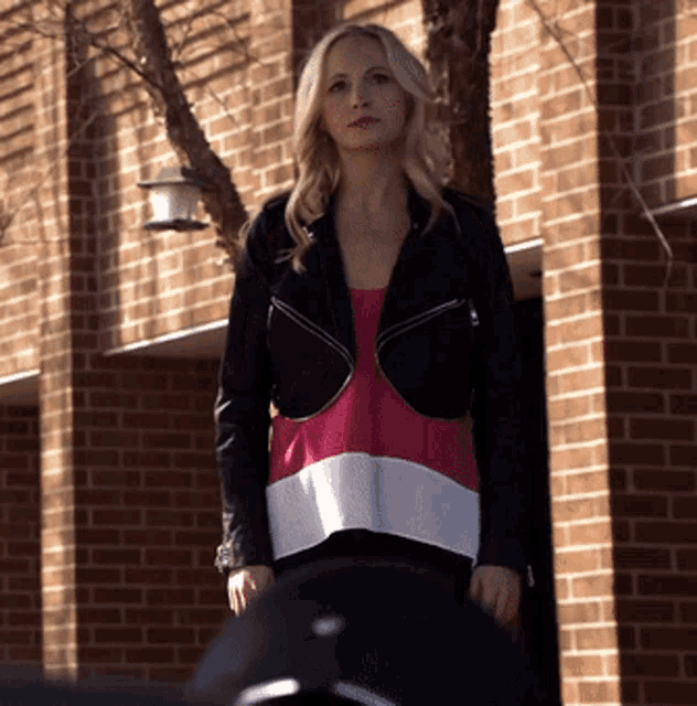 a woman standing in front of a brick building wearing a black jacket and a pink and white shirt