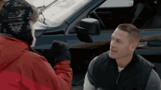a man in a red jacket is talking to another man in a black vest in front of a car