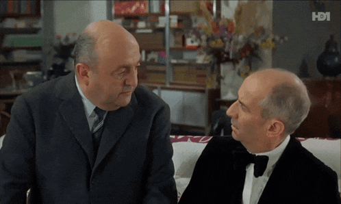 two men in suits and bow ties are sitting on a couch in front of a tv screen that says hdi