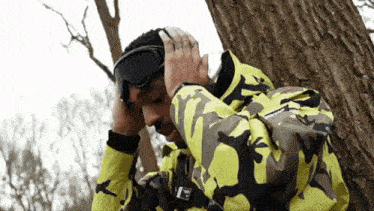a man wearing a camouflage jacket and goggles is leaning against a tree ..