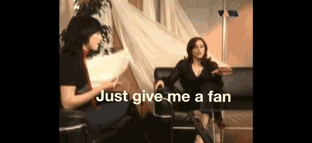 two women are sitting on a couch with the words just give me a fan behind them