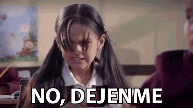 a little girl in a school uniform is sitting at a desk in a classroom and says `` no , dejenme '' .