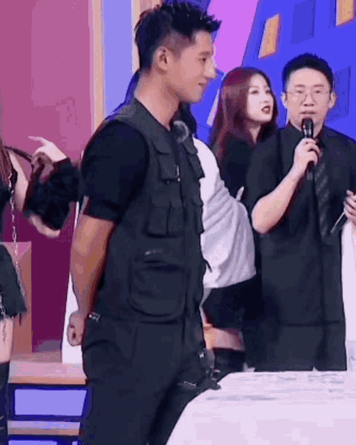 a man in a black vest is standing in front of a microphone in a room with other people .