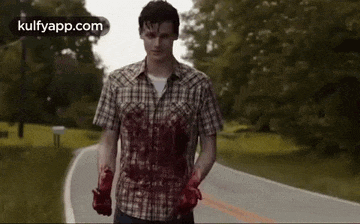a man in a plaid shirt with blood on his arms is walking down a road .