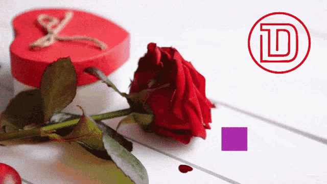 a red rose sits next to a red heart shaped box with the letter d on it