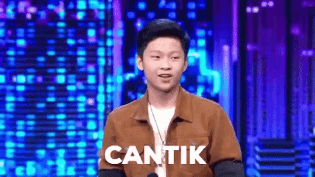 a young man in a brown jacket is standing in front of a microphone with the word cantik written on the screen behind him .