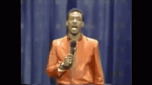 a man in an orange suit is standing on a stage holding a microphone and speaking into it .
