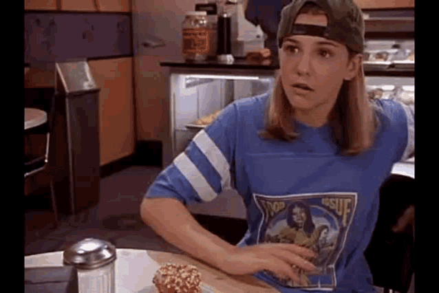 a woman wearing a baseball cap and a blue shirt is sitting at a table in a restaurant eating a hamburger .