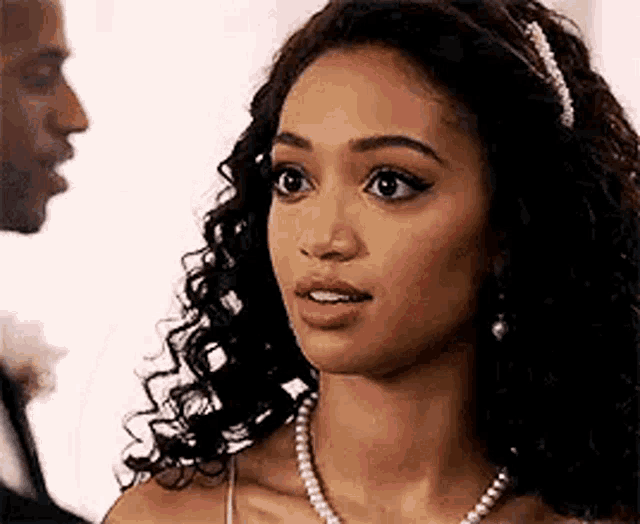 a woman with curly hair and a pearl necklace is standing next to a man in a suit .