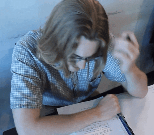 a man in a plaid shirt is sitting at a desk writing in a notebook