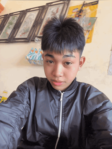 a young boy in a black jacket takes a selfie in front of a wall with pictures on it