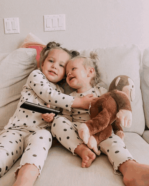 two little girls sitting on a couch with a stuffed monkey and a remote