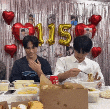 two young men are eating food in front of a wall with balloons and a number 115 balloon