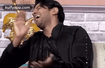 a man in a black jacket is laughing and making a funny face while sitting in front of a brick wall .