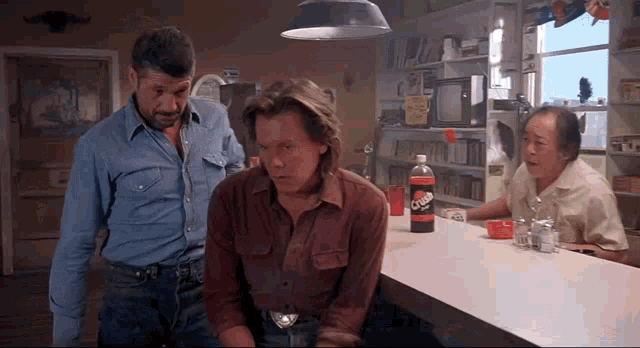 three men are standing around a counter with a bottle of crush soda