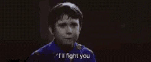 a young boy in a blue shirt is holding his fist up in the air and saying `` i 'll fight you '' .