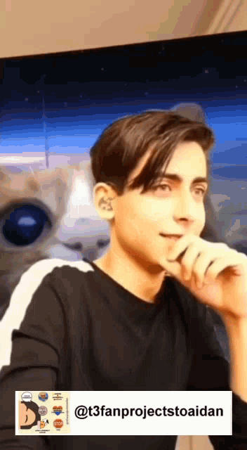 a young man in a black shirt is sitting in front of a screen with a cat behind him .