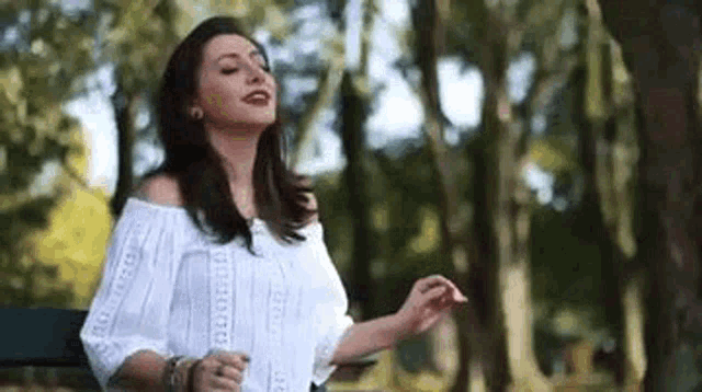 a woman in a white off the shoulder top is standing in a park with her arms outstretched .