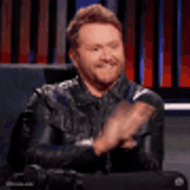 a man in a leather jacket is clapping his hands while sitting in a chair on a stage .
