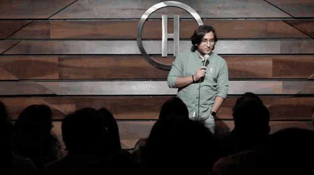 a man stands in front of a crowd holding a microphone in front of a wooden wall with the letter h on it