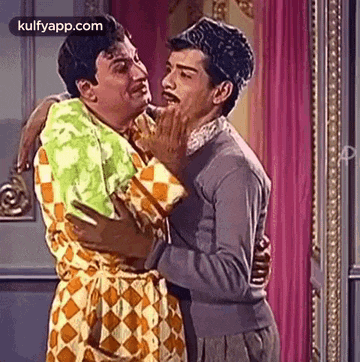 two men are hugging each other in a room and one of them is touching the other 's face .