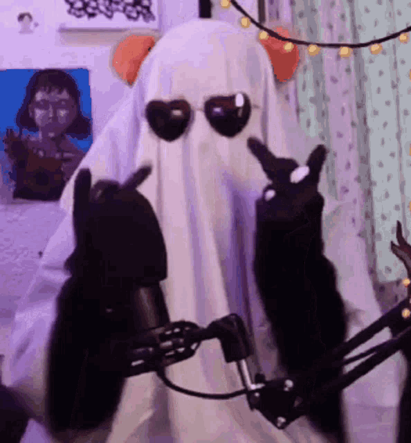 a person in a ghost costume is wearing sunglasses and giving the peace sign while sitting in front of a microphone .