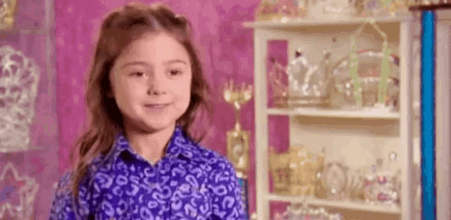 a little girl in a blue shirt is smiling in front of a shelf filled with jewelry .