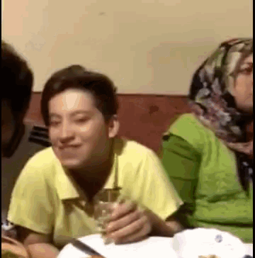 a boy in a yellow shirt is sitting at a table with a glass of wine in his hand .
