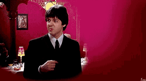 a man in a suit and tie is pointing at something while sitting at a table in front of a pink wall .