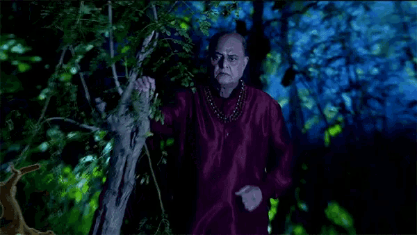 a man in a red shirt with a necklace around his neck stands in a dark forest