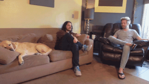 two men sit on a couch with a dog laying on it