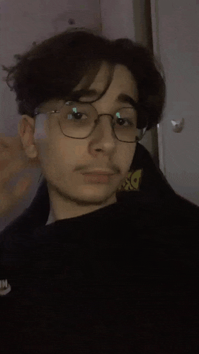 a young man wearing glasses and a black sweater is covering his face with his hand .