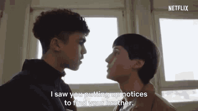 two young men are standing next to each other and talking to each other in front of a window .