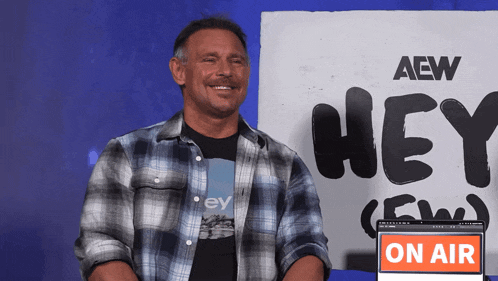 a man in a plaid shirt is smiling in front of a sign that says hey