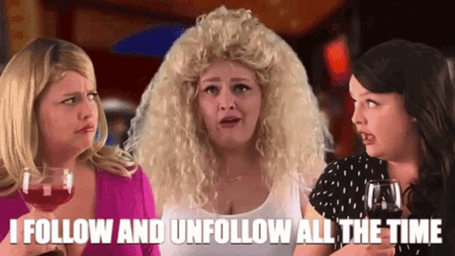 three women are standing next to each other and one of them is wearing a wig and holding a glass of red wine .