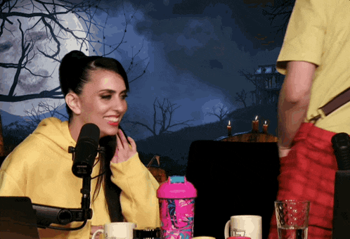a woman in a yellow hoodie smiles in front of a microphone while a man stands behind her