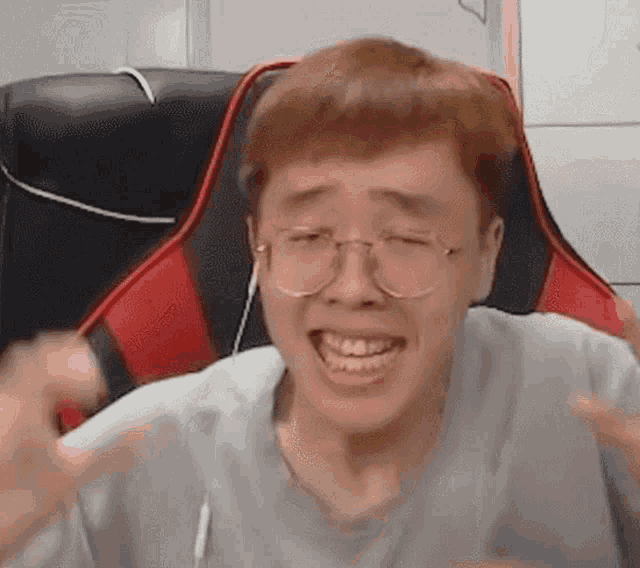 a man wearing glasses and headphones is sitting in a red and black gaming chair making a funny face .