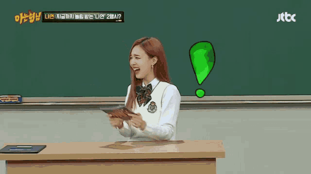 a girl in a school uniform is sitting at a desk in front of a blackboard holding a piece of paper .