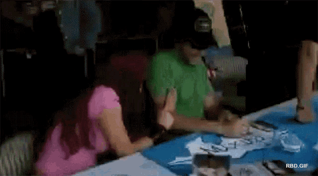 a woman in a pink shirt sits at a table with a man in a green shirt signing a piece of paper