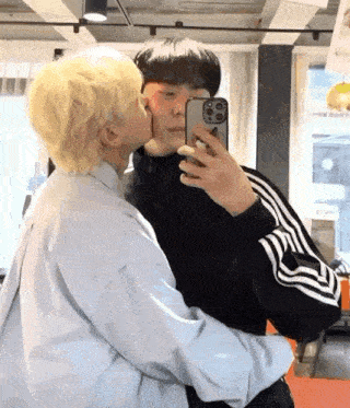 a man is kissing another man on the cheek while taking a selfie with their phone .