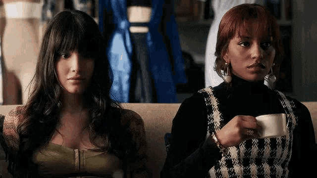 two women are sitting on a couch drinking coffee .