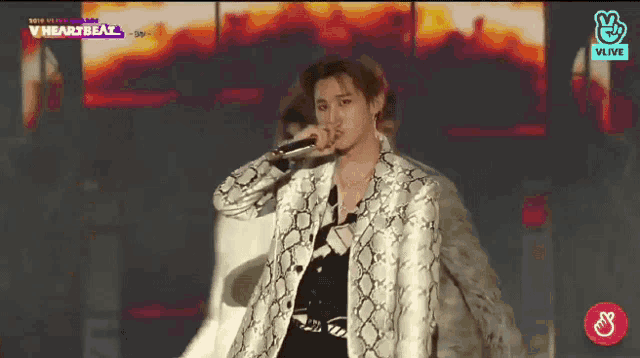 a man in a snakeskin jacket is singing into a microphone in front of a screen that says vheartbeat