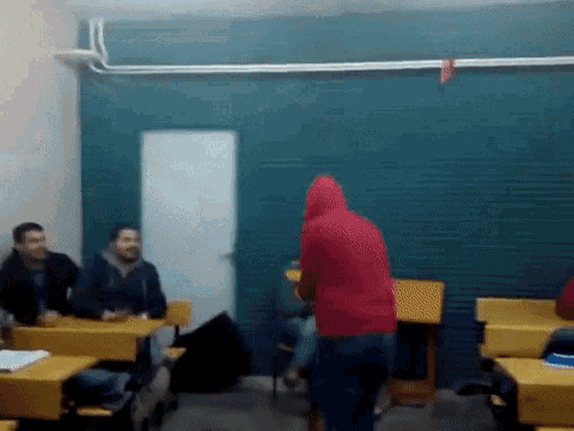 a man in a red hoodie is standing in a classroom with other people sitting at their desks .