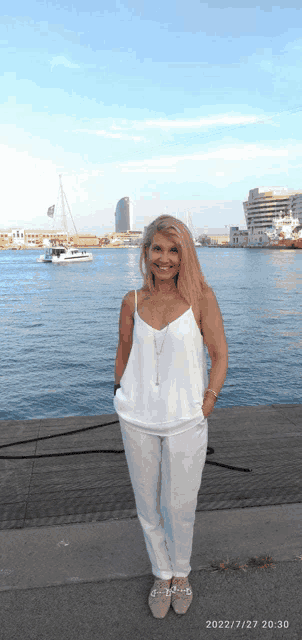 a woman in a white tank top and white pants stands in front of a body of water with a boat in the background