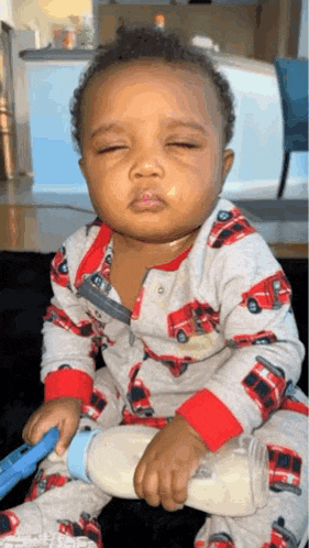 a baby in a fire truck pajamas is holding a bottle of milk and making a funny face .