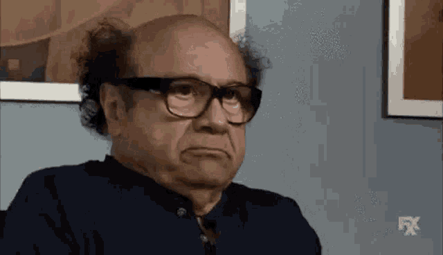 a bald man wearing glasses is making a funny face while sitting in front of a wall .